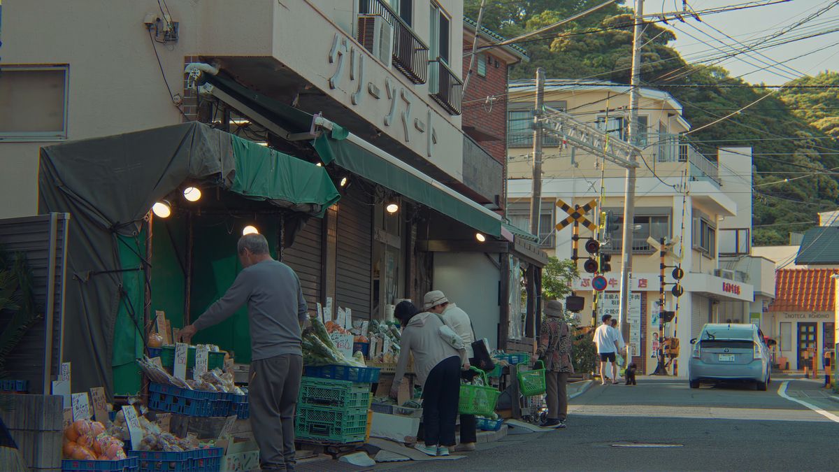 踏切のそばの八百屋の店先で野菜を選ぶ人たち