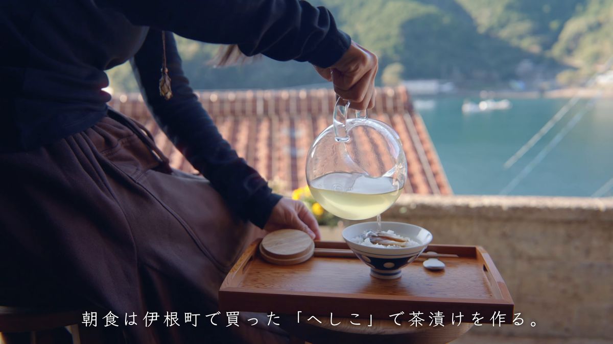 窓辺の盆の上で茶を注ぐ手元と、瓦屋根の向こうの湾