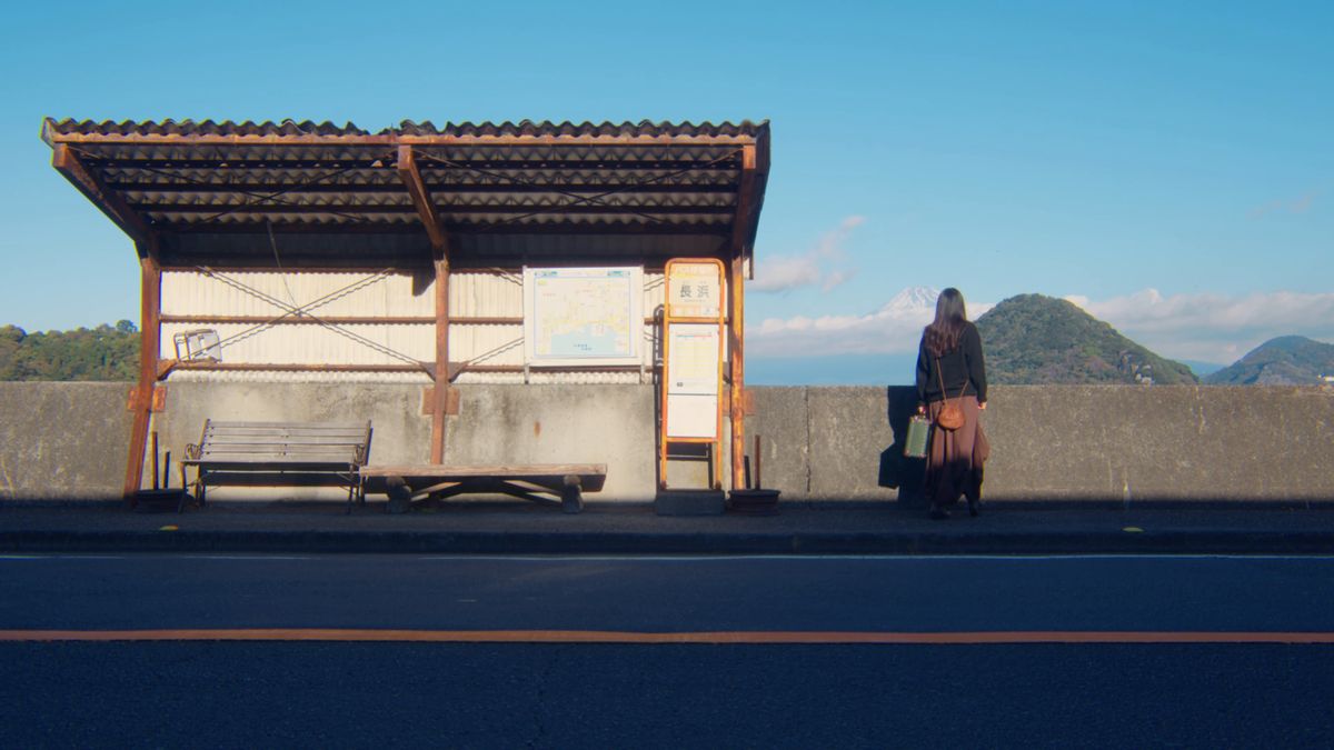 木造の屋根のバス停のベンチから立ち上がる後ろ姿と、海の向こうの山