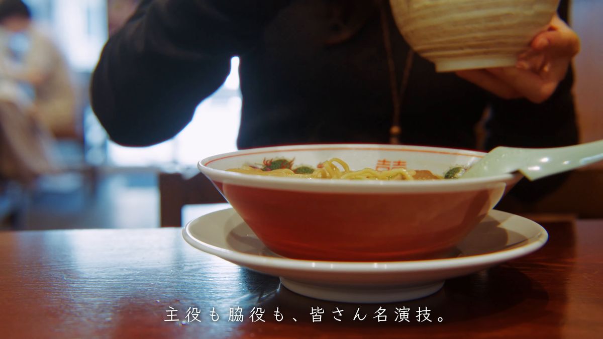 赤い丼のラーメンの奥で器を持ち上げる